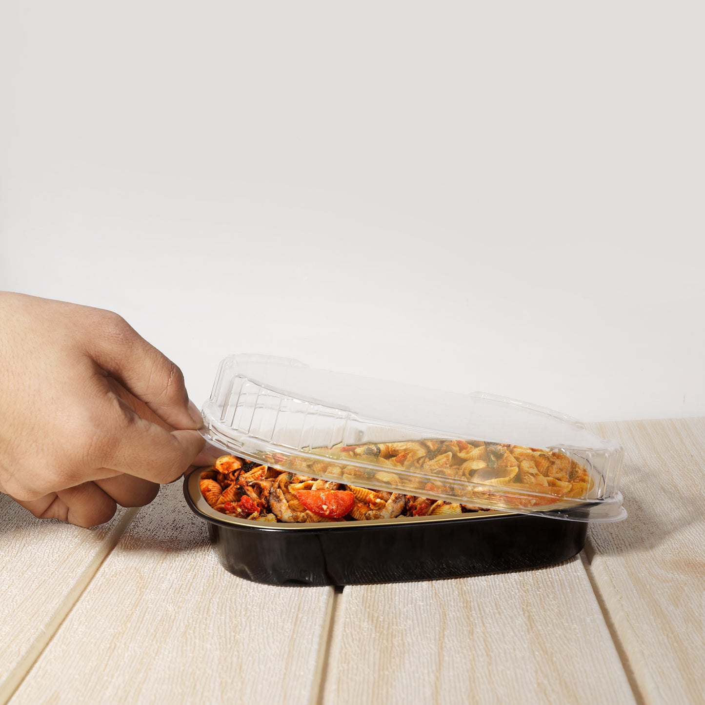 Hand lifting clear plastic lids for trays off black food container with pasta and vegetables