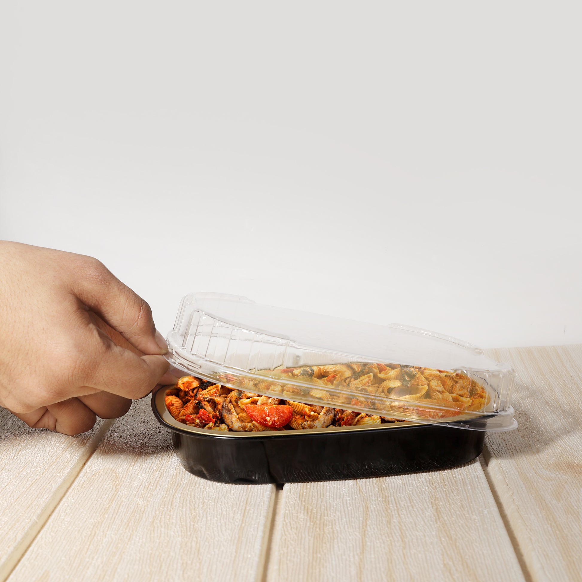 Hand lifting clear plastic lids for trays off black food container with pasta and vegetables